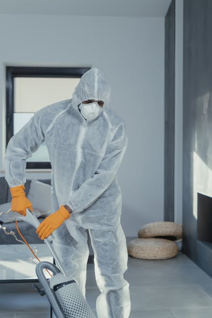 Close-up of a person wearing yellow rubber gloves spraying disinfectant from a spray bottle onto a white ceramic sink with a silver faucet, positioned in a bathroom with white tiled walls. The scene e